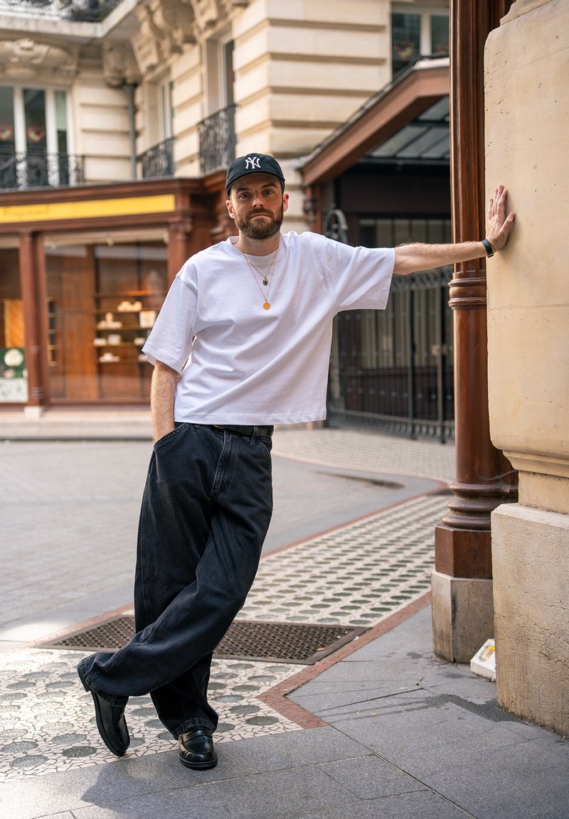 Man wearing white shirt, black jeans, black cap, and necklace leans against building wall on tiled city pavement.