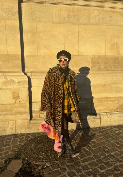 Abrigo de estampado de leopardo, camisa de cuadros amarillos, falda negra y botas con estampado. Los accesorios incluyen gafas de sol blancas y un bolso en forma de labios.