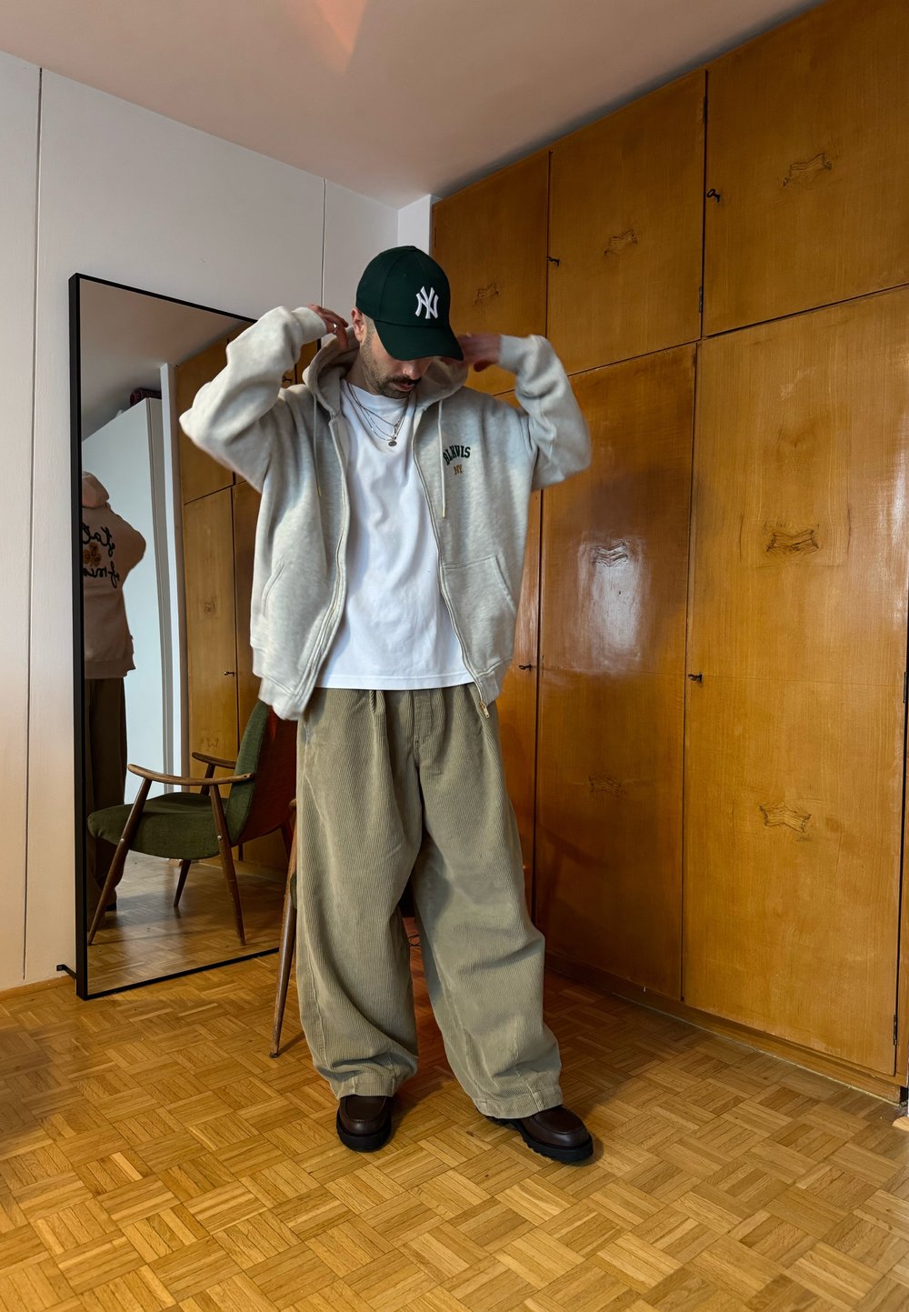 Homme ajustant son sweat à capuche, portant un t-shirt blanc, un pantalon large beige, des chaussures foncées et une casquette verte dans une pièce avec une armoire en bois et un miroir sur pied.