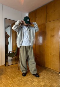 Homme ajustant son sweat à capuche, portant un t-shirt blanc, un pantalon large beige, des chaussures foncées et une casquette verte dans une pièce avec une armoire en bois et un miroir sur pied.