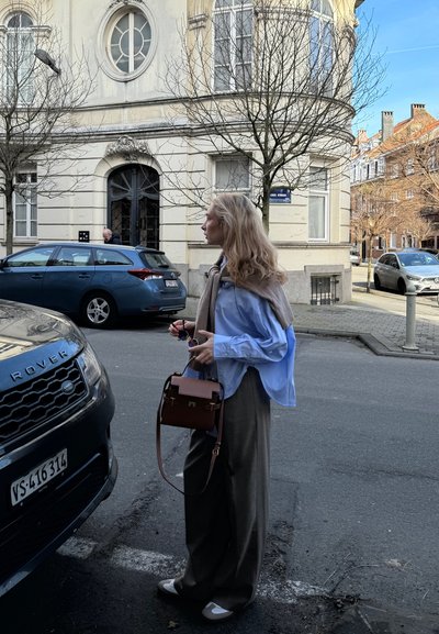 Camisa azul de gran tamaño, pantalones beiges, bolso marrón y zapatos claros. Coches aparcados y un edificio con grandes ventanas en el fondo.