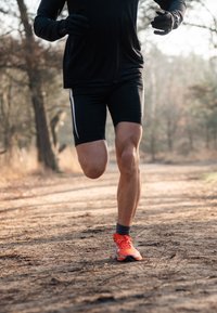 T-shirt et short de sport noirs, chaussures de course orange avec des accents noirs, posés sur un sentier en terre entouré d'arbres.