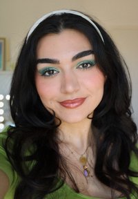 Ung kvinne med langt, bølgete mørkt hår, iført en grønn topp. Hun har dristig grønn øyeskygge og en nøytral leppestift, akkompagnert av lagdelte halskjeder.