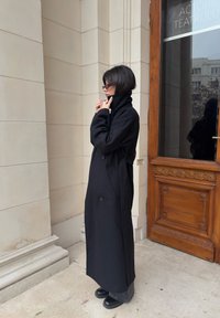 Dlhý čierny kabát vyrobený z hrubého materiálu, s vysokým golierom, dvoma gombíkmi, bočnými vreckami a opaskovým detailom v páse.