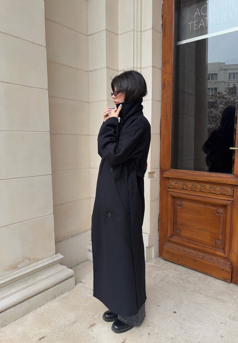 Lungo cappotto nero realizzato in un tessuto spesso, con colletto alto, due bottoni, tasche laterali e dettaglio con cintura in vita.