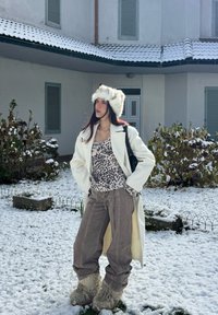 Hvid uldfrakke, leopardmønstret top, grå manchesterbukser, pelsbesat hat og pelsede støvler, sat mod en snedækket landskab med buske.