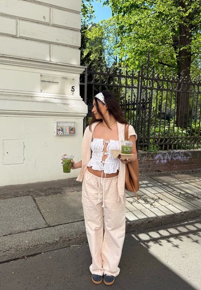 Una persona que lleva un top corto blanco con cordones, pantalones sueltos de un beige claro y un suéter ligero, sosteniendo dos bebidas y un bolso.