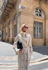 Beige trench coat nagy fekete gombokkal és övvel. Fekete kézitáska. A modellen kerek napszemüveg van. Városi utca a háttérben.