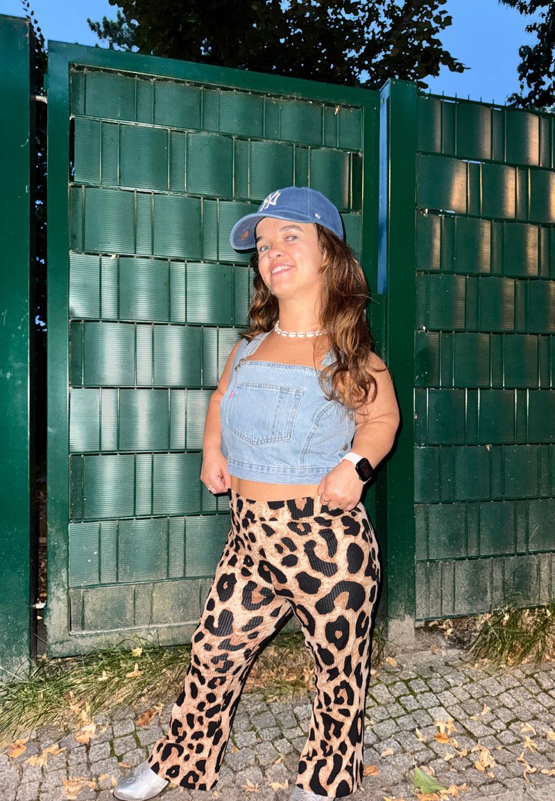 Cropped Top in denim, pantaloni larghi a stampa leopardata, sneakers argento e cappellino blu. Lo sfondo presenta un cancello verde testurizzato e pavimentazione.