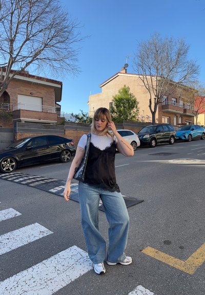 Joven mujer con cabello rubio vestida con ropa casual, de pie en un paso de peatones en un día soleado, con coches aparcados y casas en el fondo.