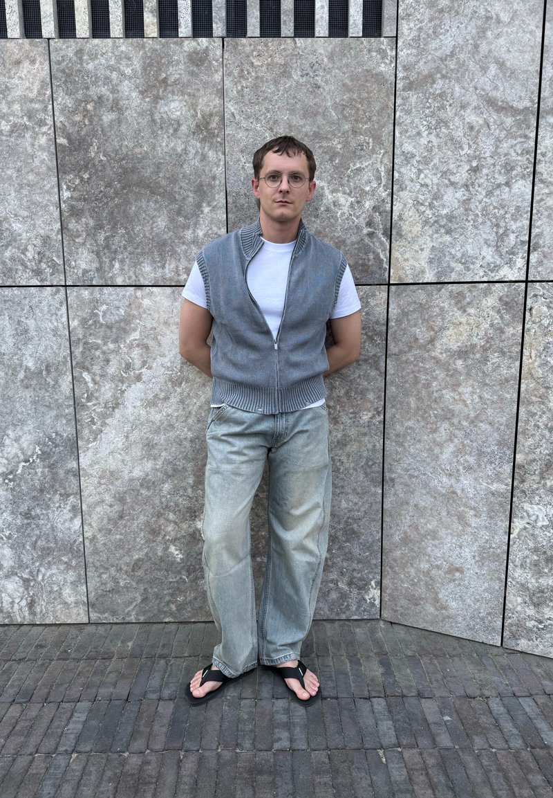 Homme avec des lunettes se tient contre un mur en pierre grise, portant un t-shirt blanc, un pull sans manches gris zippé, un jean délavé et des tongs noires.