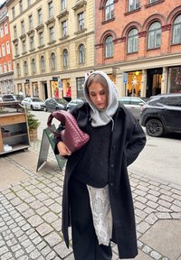 Long manteau noir par-dessus un pull noir, sweat à capuche gris texturé et détails en dentelle blanche. Porte un sac à main bordeaux tressé. Fond de rue pavée.