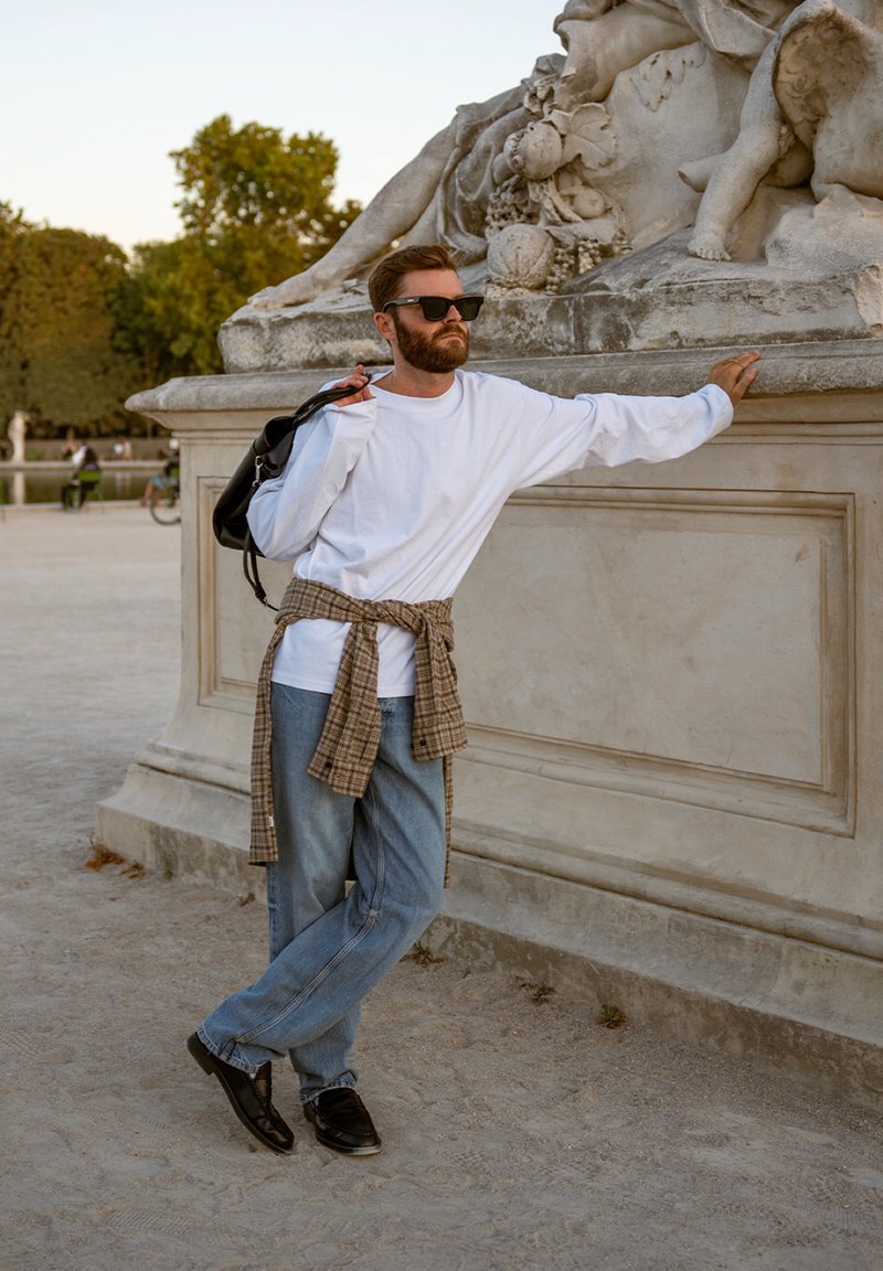 Homme portant des lunettes de soleil, une chemise blanche et un jean, appuyé contre une statue en pierre ornée dans un parc extérieur au coucher du soleil.