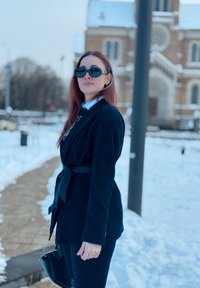 Veste noire avec ceinture nouée, portée sur une chemise bleue à col. Lunettes de soleil noires, pantalon slim et sac à main noir. Arrière-plan enneigé.