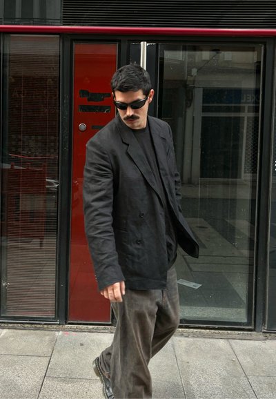 Blazer negro de gran tamaño con botones grandes, llevado sobre una camiseta negra. Pantalones oscuros de corte holgado y zapatos negros. Cabello corto y liso.