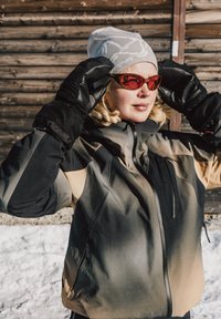 Veste de ski chaude en blocs de couleurs noir, beige et gris. Elle présente un col montant, des poches zippées, et est assortie à des gants noirs et des lunettes de soleil rouges.