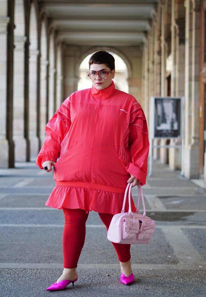 Persoon in een felrode oversized jas en leggings, met een roze handtas, staand in een gewelfde stenen corridor.