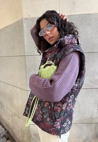 Γυναίκα με σγουρά σκουρόχρωμα μαλλιά φοράει εμπριμέ καπιτονέ γιλέκο, μωβ πλεκτό πουλόβερ, γυαλιά ηλίου, κρατώντας μικρή πλεκτή τσάντα σε ανοιχτό πράσινο χρώμα, μπροστά από πέτρινο τοίχο.