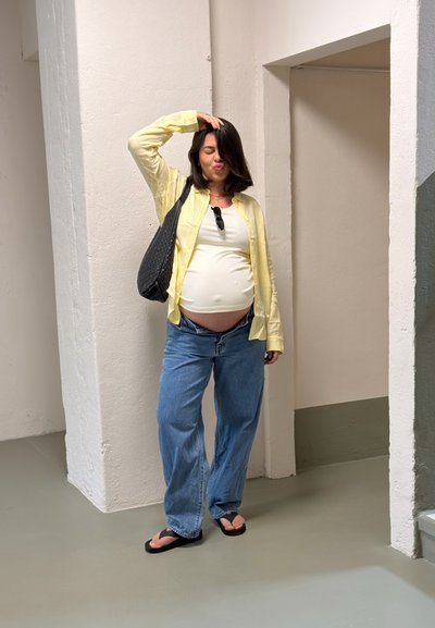 Mujer embarazada con camisa amarilla, camiseta blanca y vaqueros azules está de pie en interiores con una bolsa negra sobre el hombro, mano en la cabeza, ojos cerrados.