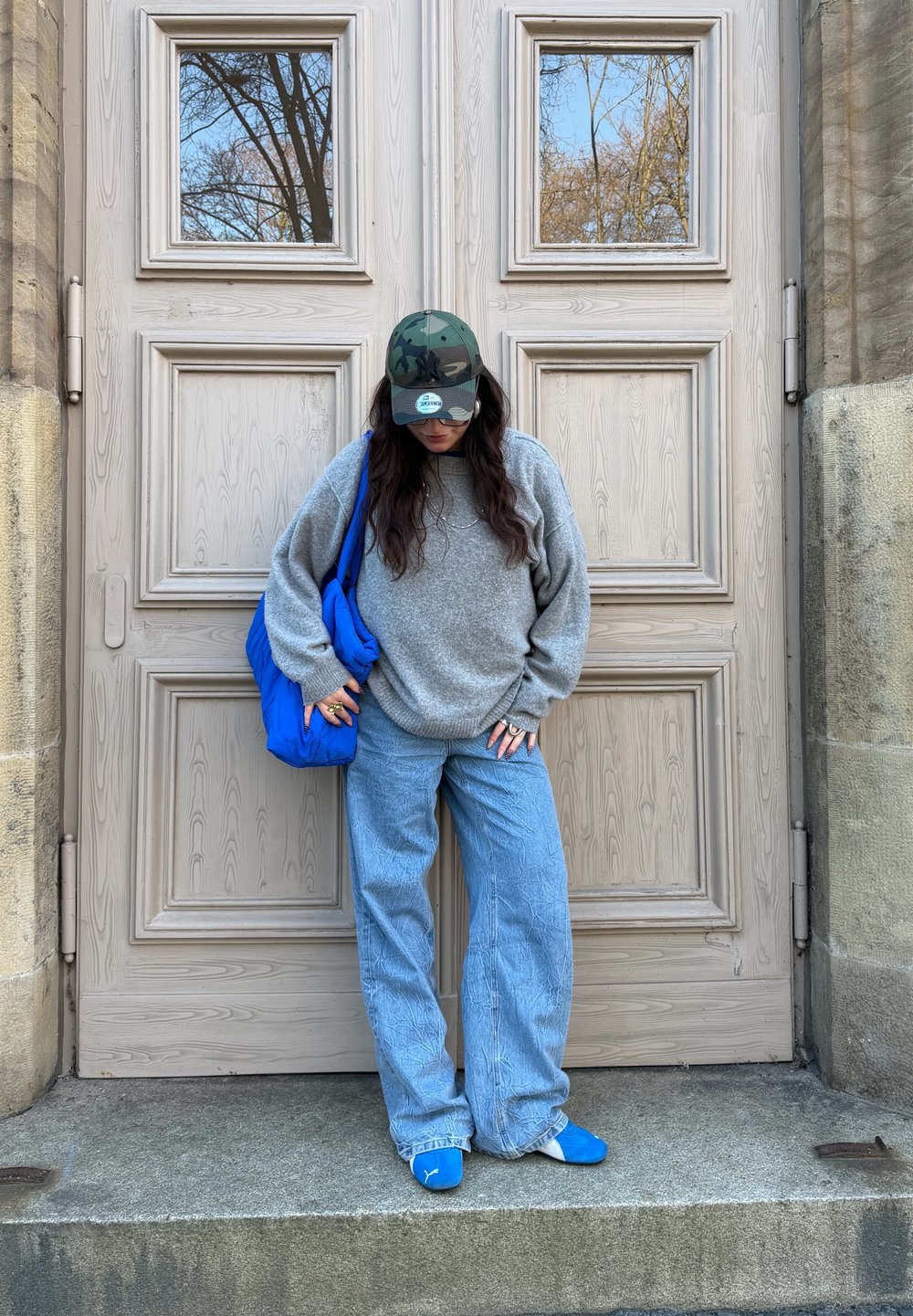 Pull gris oversized, jean pattes d\'éléphant bleu clair, sac à bandoulière bleu et casquette camouflage. Des chaussures décontractées complètent la tenue.