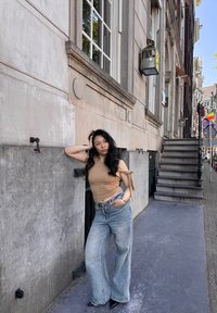Vaatteet: beige off-shoulder -paitapusero, jossa on solmut, vaaleansiniset leveälahkeiset farkut vyöllä ja teräväkärkiset mustat korkokengät, poseeraten tekstuuriseinän edessä.