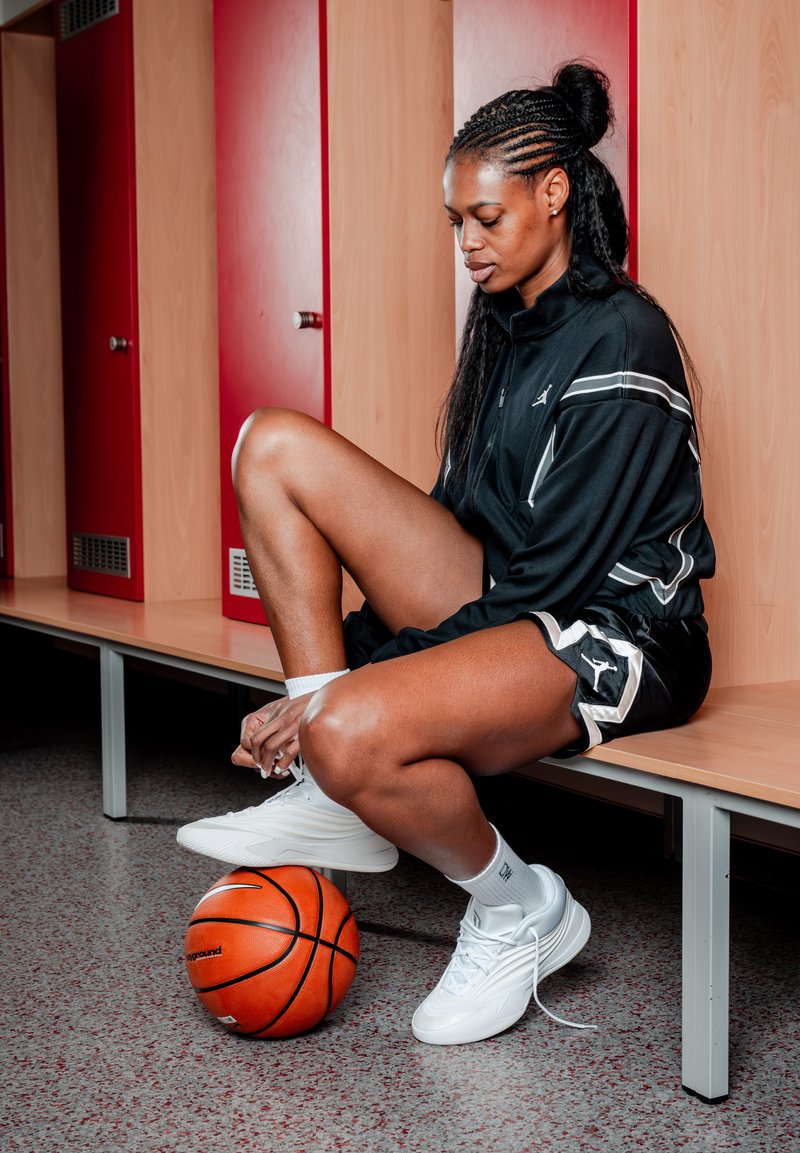 Mujer con ropa deportiva atándose los cordones en un banco en el vestuario con una pelota de baloncesto bajo el pie, rodeada de taquillas rojas y de madera.