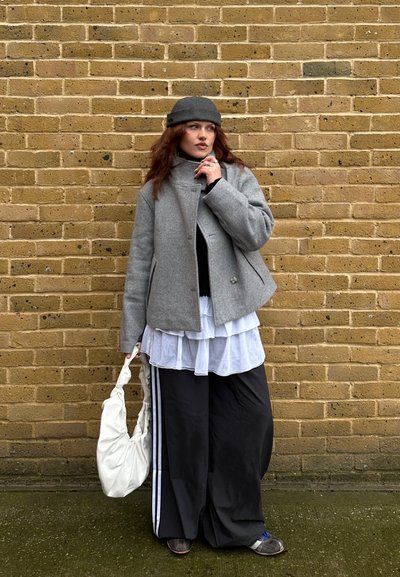 Mujer con abrigo y sombrero grises, blusa blanca de varias capas, pantalones anchos negros con rayas blancas, sosteniendo una bolsa blanca, de pie frente a una pared de ladrillos amarillos.