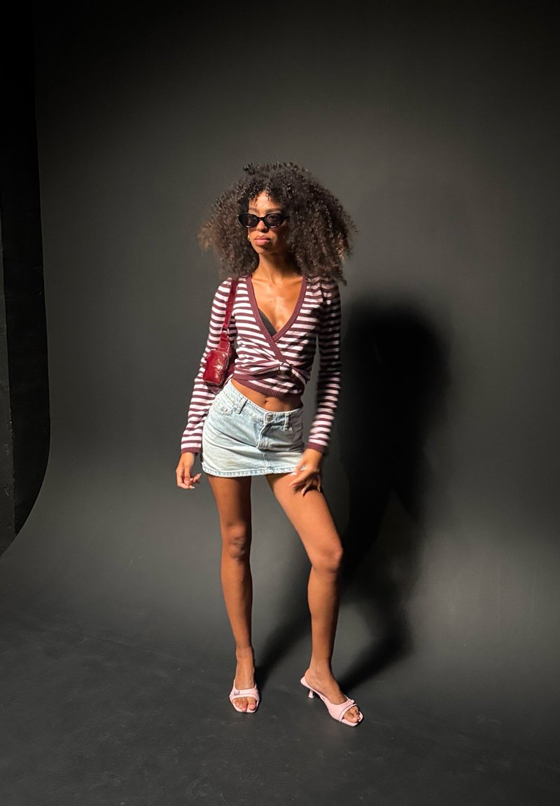 Femme aux cheveux bouclés portant des lunettes de soleil, un top cache-cœur rayé, une mini jupe en denim, des talons roses et un sac à épaule rouge, debout devant un fond sombre.