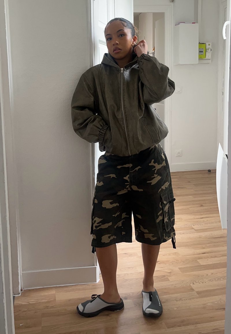 Jeune femme à l'intérieur portant une veste zippée verte, un short cargo camouflage et des chaussures slip-on noires et blanches, debout sur un sol en bois.