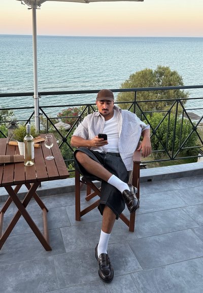 Un hombre con una camisa a rayas y una gorra marrón está sentado en una silla de madera, sosteniendo un teléfono, con una botella de vino y copas sobre una mesa, con el océano de fondo.