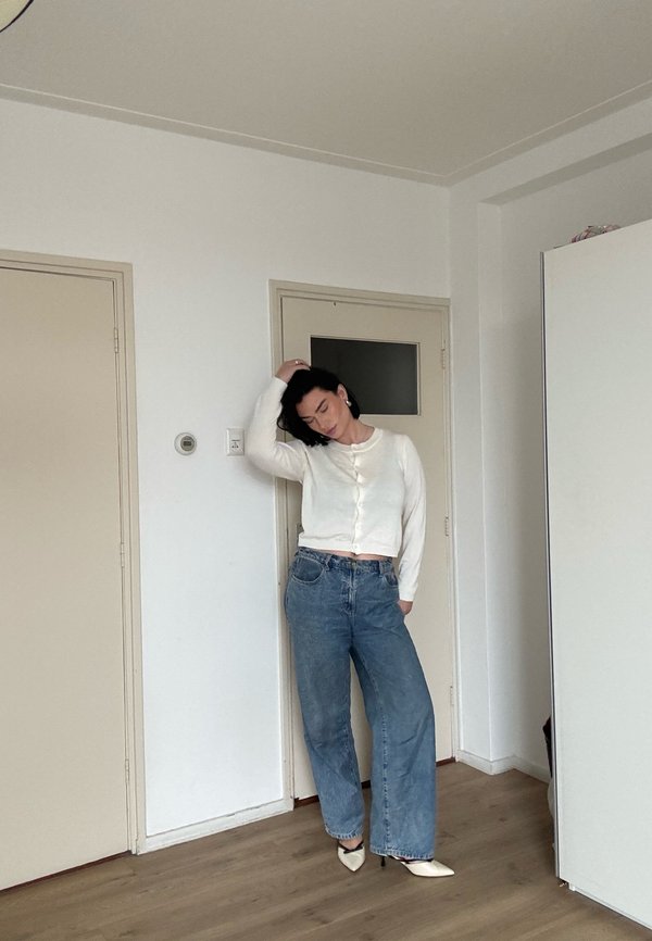 Kvinne som står innendørs iført hvit knappet cardigan, vide blå jeans og hvite spisse hæler, med en hånd på hodet og øynene lukket.