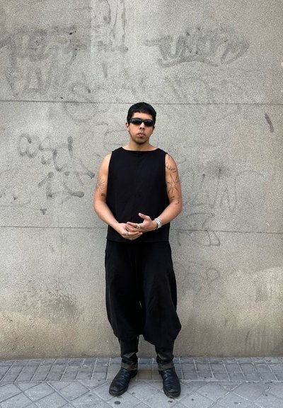 Top negro sin mangas y pantalones sueltos negros de tela texturizada. Los accesorios incluyen anillos y un reloj. Se llevan gafas de sol oscuras.