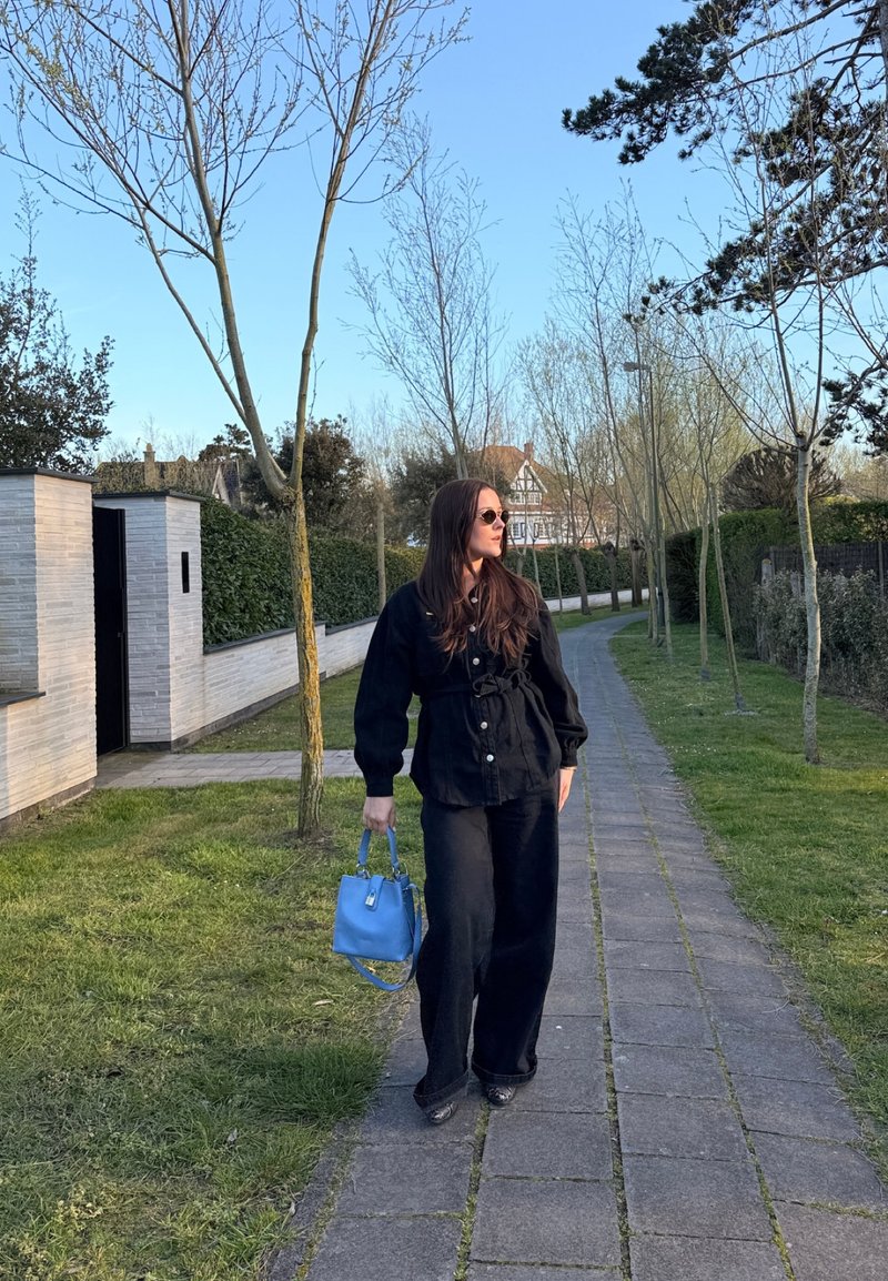 Woman in black outfit and sunglasses walking on paved path, holding a blue handbag, with leafless trees and houses in the background.