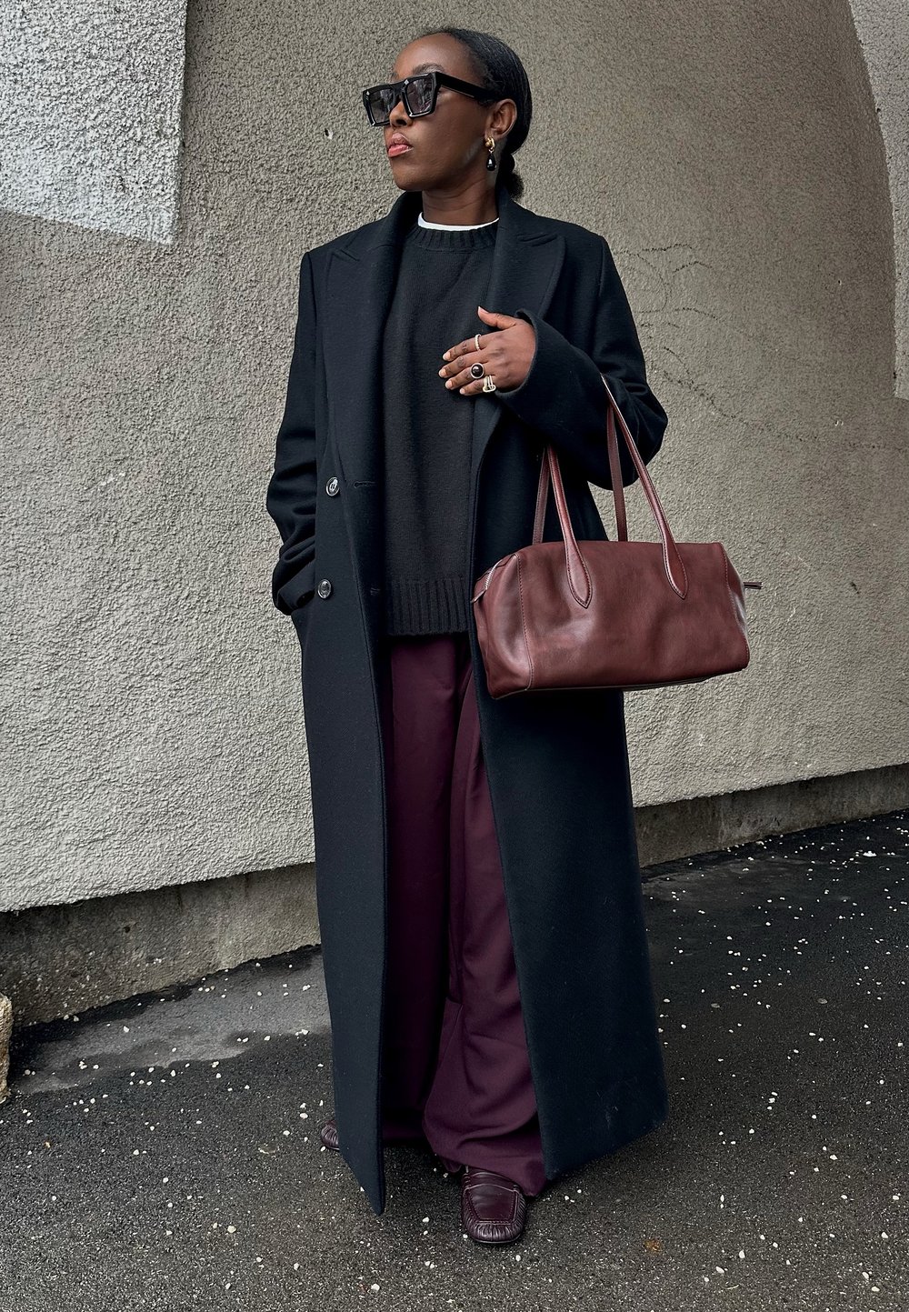 Γυναίκα που φορά μαύρο μακρύ παλτό, σκούρο πουλόβερ, φαρδιά μωβ παντελόνια, μεγάλα γυαλιά ηλίου, κρατώντας καφέ σκούρα δερμάτινη τσάντα, με φόντο τοιχο με υφή.