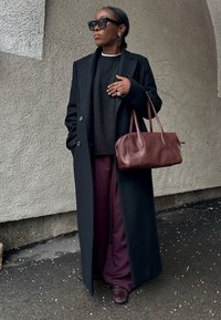 Γυναίκα που φορά μαύρο μακρύ παλτό, σκούρο πουλόβερ, φαρδιά μωβ παντελόνια, μεγάλα γυαλιά ηλίου, κρατώντας καφέ σκούρα δερμάτινη τσάντα, με φόντο τοιχο με υφή.