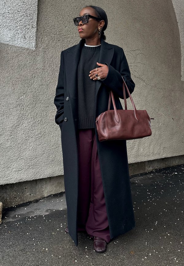 Frau trägt schwarzen langen Mantel, dunklen Pullover, violette weite Hose, große Sonnenbrille, hält eine dunkelbraune Lederhandtasche vor einem strukturierten Wandhintergrund.