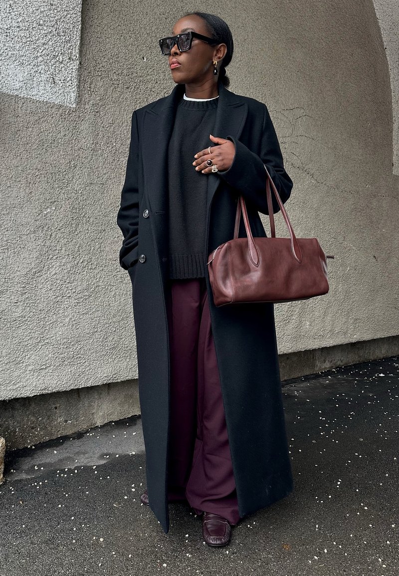 Kvinde iført sort lang frakke, mørk sweater, lilla vide bukser, store solbriller, holder mørkebrunt læderhåndtaske foran en væg med tekstur.