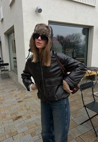 Femme portant un chapeau en fourrure, une veste en cuir noire et des lunettes de soleil, debout à l'extérieur d'un café avec une terrasse sur un trottoir carrelé.