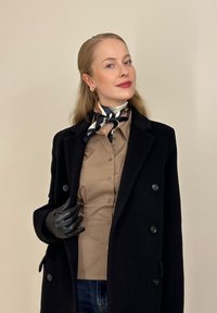 Γυναίκα που φορά μαύρο παλτό, καφέ πουκάμισο με κουμπιά, μαύρο δερμάτινο γάντι, μαντήλι με σχέδια στο λαιμό και χρυσά σκουλαρίκια με φόντο μπεζ.