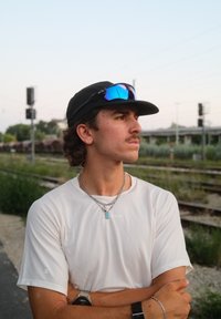 Fehér, rövid ujjú ing, ezüst lánc nyaklánc medállal, fekete sapka kék tükörszemüveggel, áll a vonatsínek mellett, növényzettel.