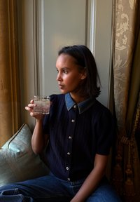 Cardigan en maille marine avec un col en denim, orné de boutons dorés. Assis sur un coussin, tenant un verre en cristal avec une boisson.