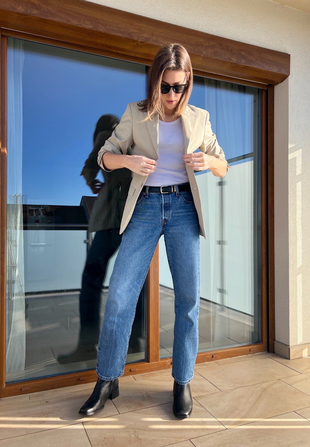 Blazer bege sobre uma blusa branca, combinado com jeans de perna reta azul claro e botas de cano curto pretas, num fundo de vidro.