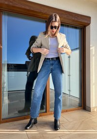 Blazer bege sobre uma blusa branca, combinado com jeans de perna reta azul claro e botas de cano curto pretas, num fundo de vidro.