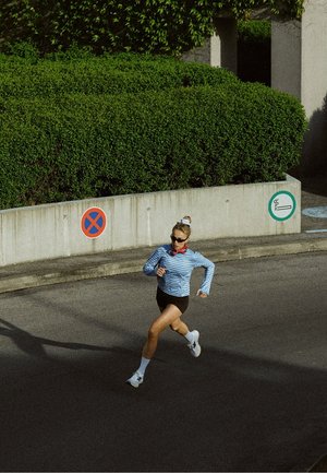 Mujer con gafas de sol y ropa deportiva corriendo por una carretera pavimentada cerca de barreras de hormigón y arbustos verdes durante el día.