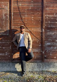 Homme portant des lunettes de soleil, un blazer beige, une chemise blanche et une casquette, appuyé contre un mur en bois usé par le temps au soleil, une jambe pliée, mains détendues.