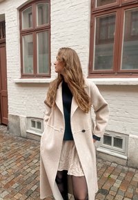 Moteris su ilgais plaukais, vilkinti smėlio spalvos paltą, juodą palaidinę ir sijoną, stovinti ant akmenimis grįstos gatvės priešais baltų plytų pastatą.