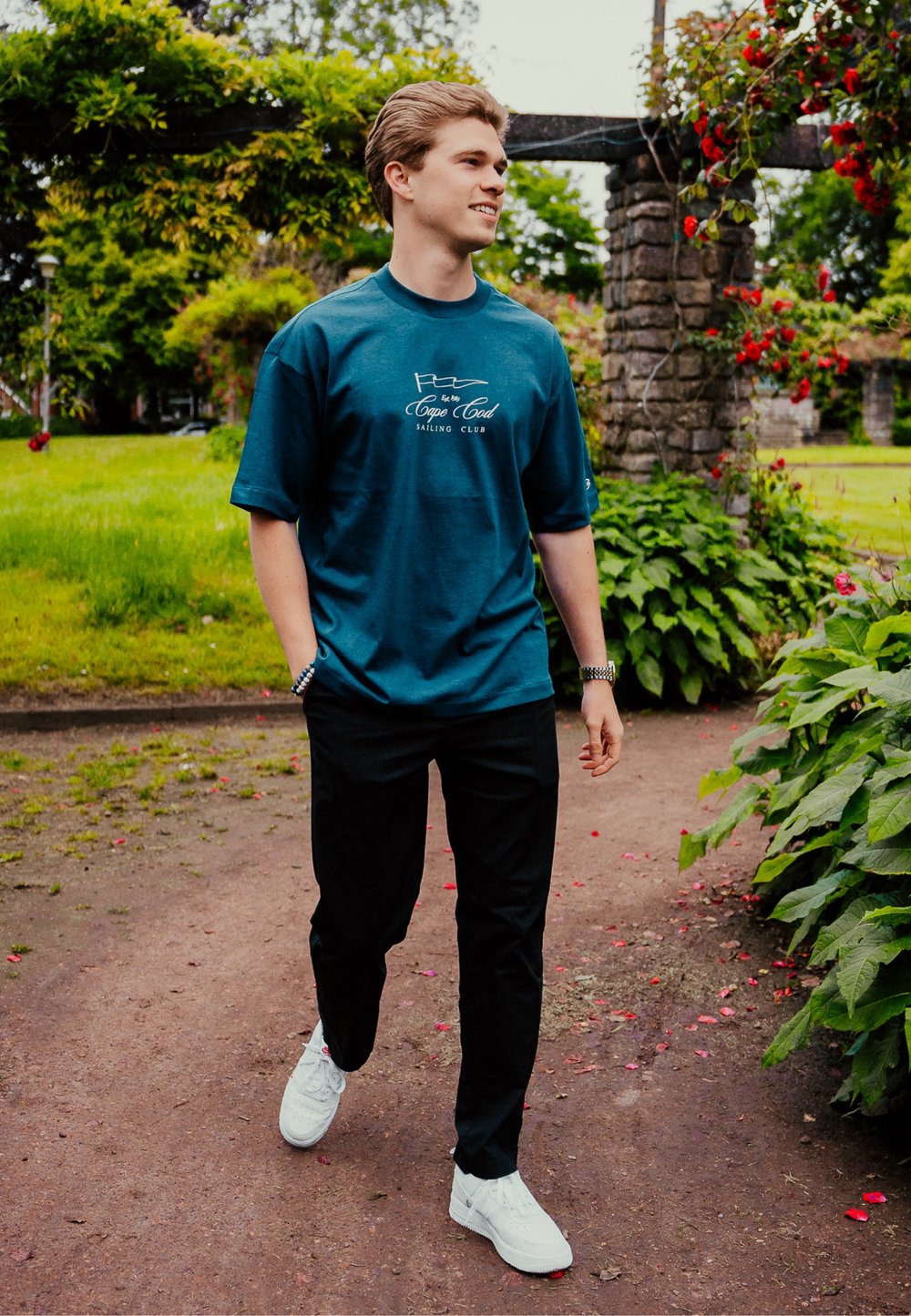 Turkusowa bawełniana koszulka z krótkim rękawem, z białym logo klubu żeglarskiego, w połączeniu z czarnymi spodniami i białymi trampkami, spacerująca po ścieżce.