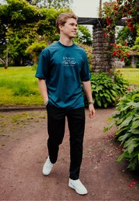 Turkusowa bawełniana koszulka z krótkim rękawem, z białym logo klubu żeglarskiego, w połączeniu z czarnymi spodniami i białymi trampkami, spacerująca po ścieżce.