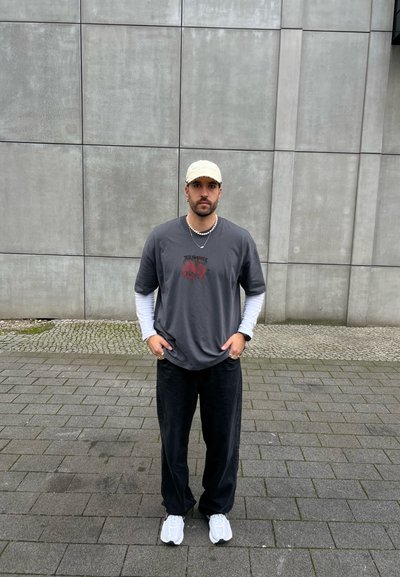 Camiseta gris de gran tamaño con estampado gráfico rojo, superpuesta con una camisa de manga larga blanca, combinada con pantalones negros y zapatillas blancas, llevando una gorra beige.