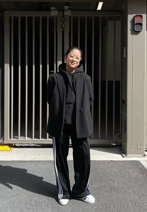 Blazer negro sobre una sudadera negra, combinado con pantalones de chándal negros con rayas blancas. Zapatillas blancas. El fondo muestra un área cerrada.